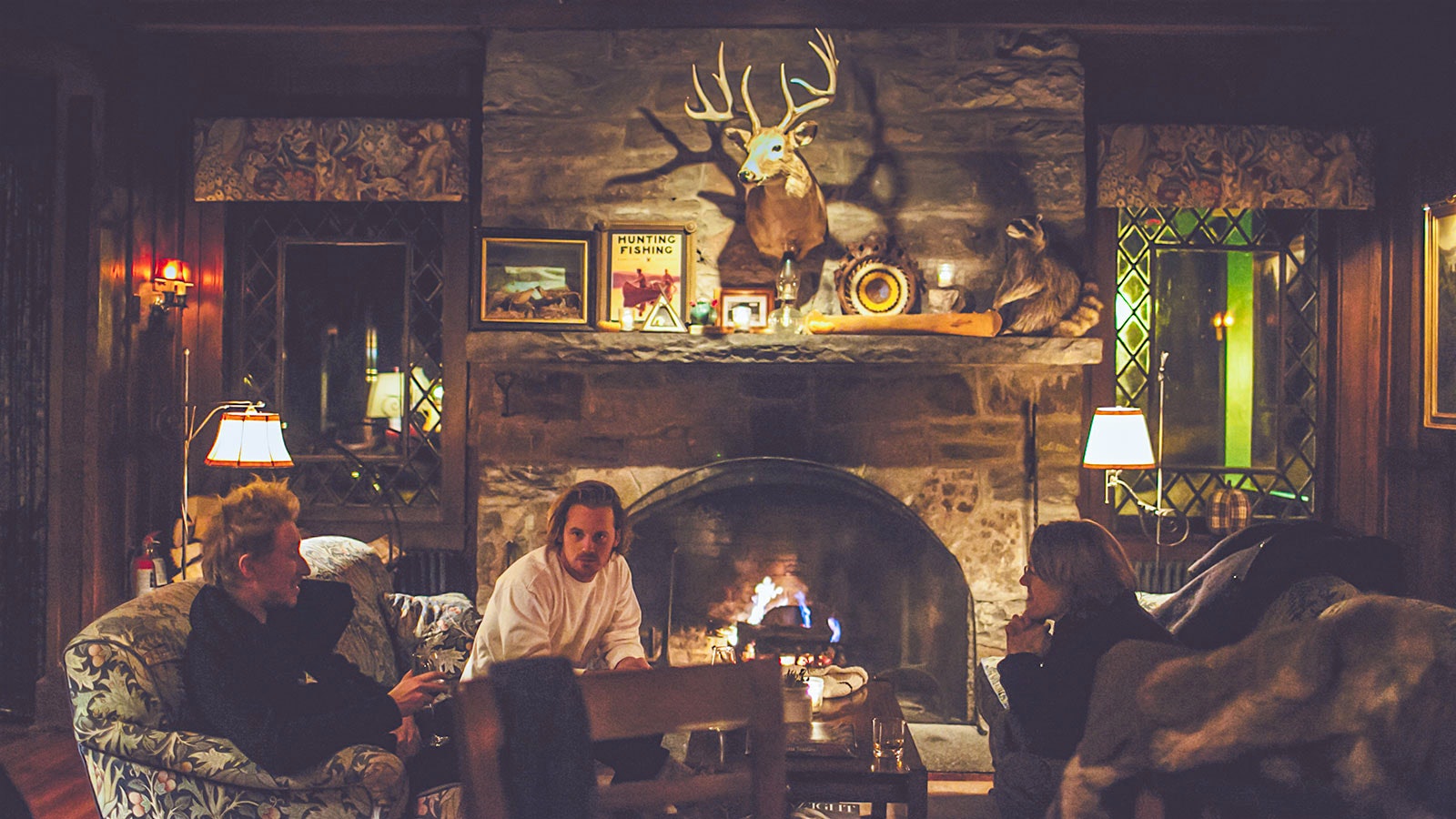 Three people sitting on couches at Deer Mountain Inn, in front of a stone fireplace and a deer trophy hanging over it
